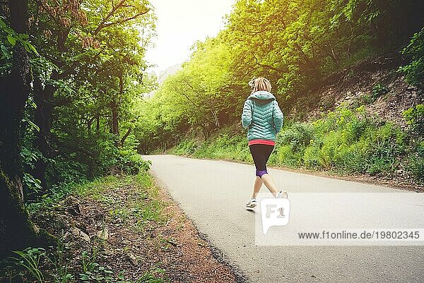 Junge Fitness blonde Frau mit Kopfhörern läuft am Morgen kaukasischen Waldweg in Sonne Licht. Seitenansicht von hinten