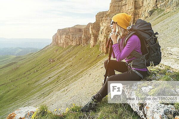Lächelnde Reisende mit gelbem Hut und Sonnenbrille sitzt am Fuße eines epischen Felsens mit einem Rucksack daneben und schaut weg