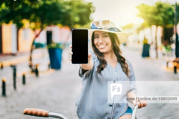 Lächelnder Tourist auf dem Fahrrad zeigt Handybildschirm auf der Straße. Schönes Mädchen mit Hut auf dem Fahrrad zeigt Handybildschirm auf der Straße