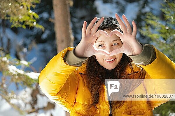 Attraktive kaukasische junge Frau zeigt Herz Geste mit Händen und schauen durch sie lächelnd im Winterwald. Ein Mädchen in warmer Winterkleidung wünscht allen Liebe