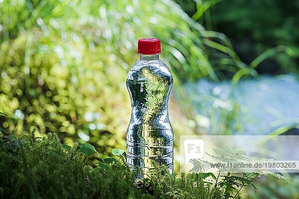 Transparenter Kunststoff Eine Flasche mit sauberem Wasser mit rotem Deckel steht im Gras und Moos vor dem Hintergrund eines zerklüfteten Bergflusses. Das Konzept des reinen natürlichen Trinkwassers
