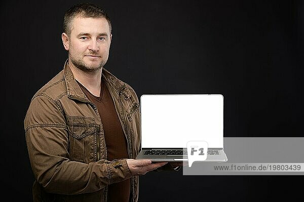 Attraktiver  positiver  freundlicher  kaukasischer Männchen  der einen Laptop in den Händen hält und seinen ausgeschnittenen Bildschirm mit Kopierbereich auf schwarzem Hintergrund präsentiert. Studioaufnahme