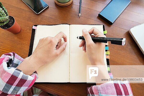Junge kaukasische attraktive Frau macht sich Notizen in ihrem Notizbuch  während sie in der Küche am Tisch sitzt. Papier Notebook schreiben weibliche Hände