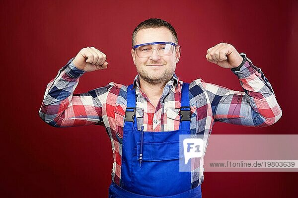 Porträt eines positiven  freundlichen kaukasischen männlichen Vorarbeiters in Latzhose und Schutzbrille auf einem dunkelroten Hintergrund. Er lächelt und schaut in die Kamera. Studioaufnahme. Zeigt eine Geste der Stärke