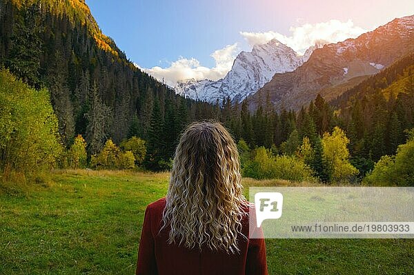 Rückansicht  eine blonde Frau in einem roten Mantel steht vor der Kulisse schneebedeckter Berge und eines dichten grünen Waldes