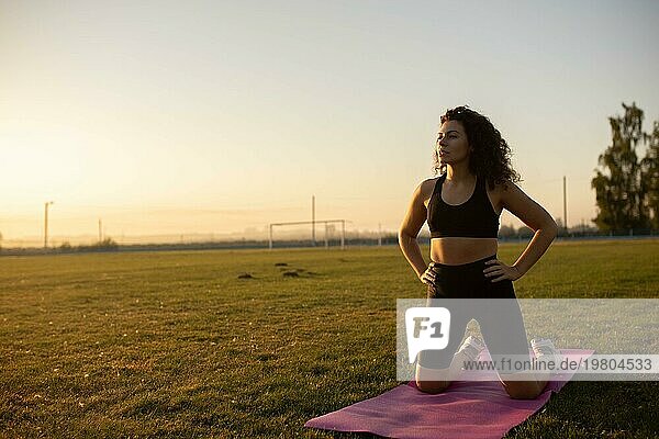 Junges lockiges sportliches Mädchen in Sportkleidung macht Yoga auf einer Yogamatte im Freien auf dem Rasen bei Sonnenuntergang