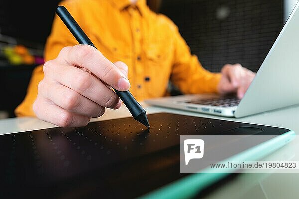 Lässiger Mann  Freiberufler mit Stift  schreibt auf digitalem Tablet Bildschirm  arbeitet an Laptopcomputer im Büro Schreibtisch. Grafik oder Webdesigner bei der Arbeit an seinem Web Design Projekt  Nahaufnahme