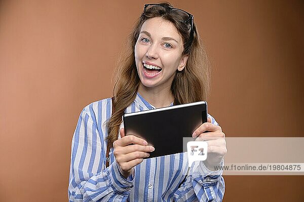 Porträt eines fröhlichen  charmanten Mädchens  das ein digitales Tablet mit einem breiten Lächeln hält  mit Hemd und Brille  Studioaufnahme