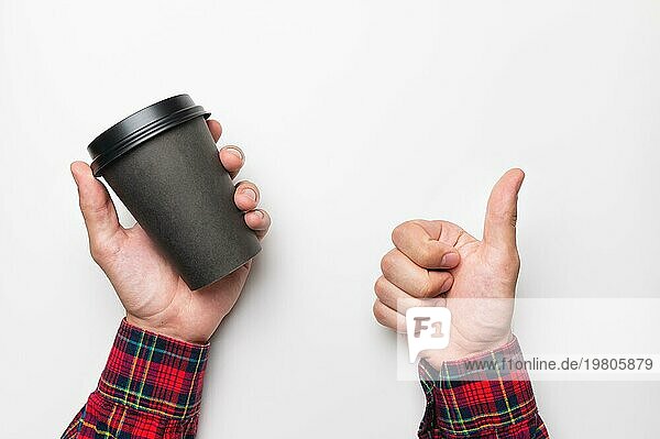Die Hand eines Mannes hält eine leere schwarze Papiertasse mit Kaffee auf weißem Hintergrund in einer Hand und zeigt mit der anderen Hand eine Geste der Klasse  Daumen hoch