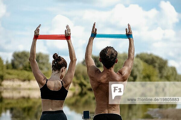 Junges sportliches Paar  das an einem sonnigen Tag am Ufer eines Sees Übungen mit einem Widerstandsband macht