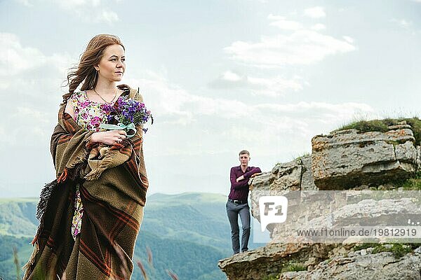 Porträt einer jungen Braut in einem bunten Kleid  eingewickelt in eine Decke und mit einem Strauß Wildblumen in der Natur  die auf dem Hintergrund ihres Freundes steht