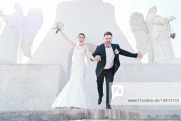 Porträt eines schönen Paares von Jungvermählten an einem Hochzeitstag mit einem Blumenstrauß in den Händen laufen die Treppe hinunter lachend und lächelnd vor dem Hintergrund eines orthodoxen christlichen Denkmal mit Engeln. Das Konzept der christlichen Hochzeit in der Kirche des
