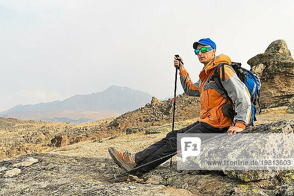 Ein einsamer Tourist mit einem Rucksack und Stöcke für die Bewegung in den Bergen. In Sonnenbrille und mit einem Rucksack Rest sidiya auf einem Stein
