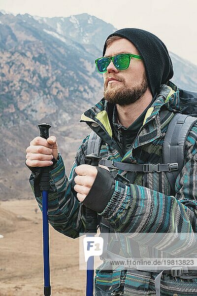 Ein Mann mit Bart und Sonnenbrille in einer Membranjacke  Hut  mit einem Rucksack und Stöcken für Nordic Walking  ein Reisender  der in der freien Natur steht und auf die Berge schaut