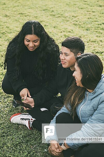 Drei Personen hispanisch lateinamerikanischer Ethnizität sitzen auf dem üppigen grünen Gras eines Parks  mit einem Smartgerät. Mit entspannten und lächelnden Gesichtsausdrücken genießen sie einen Moment der Muße inmitten der Hektik des Alltags