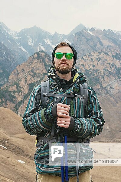 Ein Mann mit Bart und Sonnenbrille in einer Membranjacke  Hut  mit einem Rucksack und Stöcken für Nordic Walking  ein Reisender  der in der freien Natur steht und auf die Berge schaut