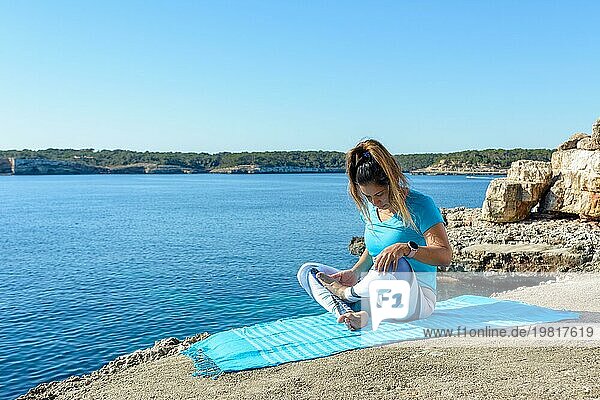 Eine Frau sitzt an einem sonnigen Tag auf einer Yogamatte am Meer  umgeben von Felsen und dem ruhigen Wasser