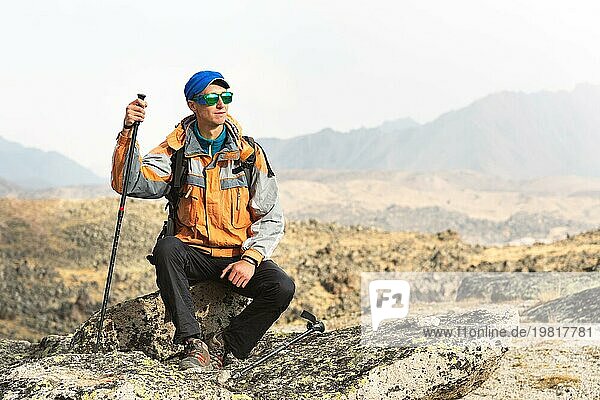 Ein einsamer Tourist mit einem Rucksack und Stöcke für die Bewegung in den Bergen. In Sonnenbrille und mit einem Rucksack Rest sidiya auf einem Stein
