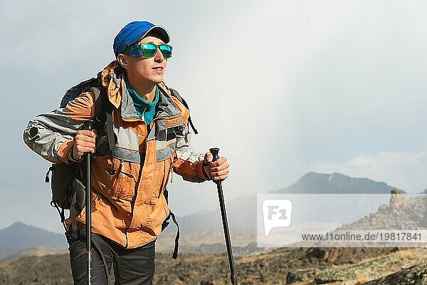 Ein einsamer Tourist mit Rucksack und Sonnenbrille genießt die Aussicht hoch in den Bergen des Kaukasus  wo es kein Gras gibt  das Dorf und den Schnee bei Sonnenuntergang
