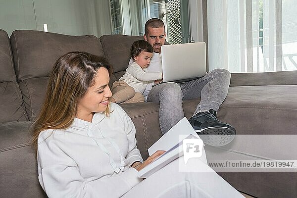 Die Mutter liest ein Buch  während der Vater auf dem Sofa sitzend einen Laptop benutzt  während der Wochenendaktivitäten einer Familie mit einem Kind zu Hause