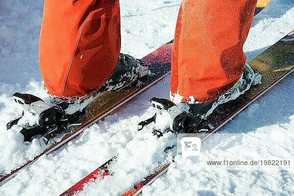 Orangefarbene Alpinskischuhe in einer Skihalterung. Ein Schuh ist vollständig auf den Skiern befestigt  der zweite nicht. Nahaufnahme. Orangefarbener Anzug