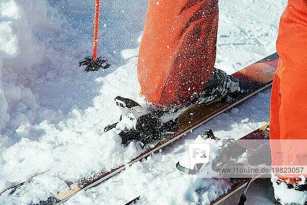 Orangefarbene Alpinskischuhe in einer Skihalterung. Ein Schuh ist vollständig auf den Skiern befestigt  der zweite nicht. Nahaufnahme. Orangefarbener Anzug