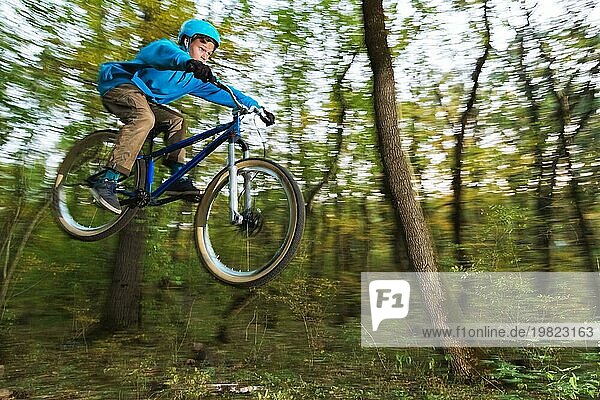 Ein junger Fahrer mit Helm und blauem Sweatshirt fliegt auf einem Fahrrad  nachdem er von einem hohen Kicker auf einem Waldradweg gesprungen ist. Aufnahme mit Langzeitbelichtung Verdrahtung