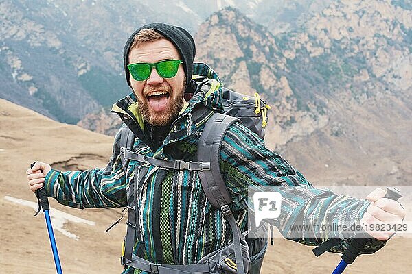 Ein Typ  der aussieht wie ein Hipster  zeigt seine Zunge und schreit in den Bergen  streckt seine Hände in verschiedene Richtungen aus und hält einen Stock für den skandinavischen Spaziergang