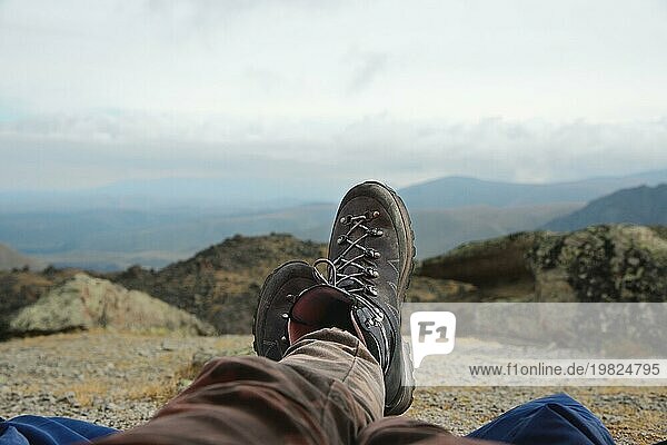 Beine eines Mannes  der sich in Stiefeln für eine Bergwanderung ausruht  vor dem Hintergrund von Bergen und Tälern mit lärmenden Wolken Wanderstiefel in den Bergen. Zeit zum Ausruhen. Freiheit Konzept