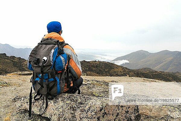 Ein einsamer Tourist mit einem Rucksack und Stöcke für die Bewegung in den Bergen. In Sonnenbrille und mit einem Rucksack Rest sidiya auf einem Stein. Blick von hinten