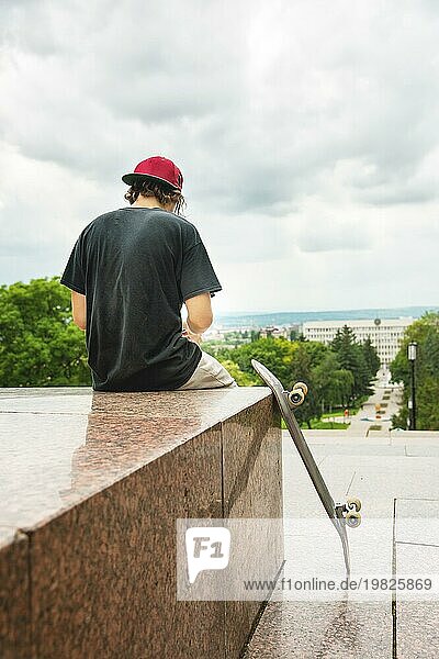 Der langhaarige Skater mit Mütze und TShirt sitzt mit dem Rücken zur Kamera und denkt neben dem Skateboard vor dem Hintergrund der Landschaft