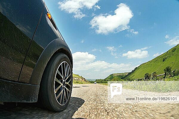 Eine Nahaufnahme der Seite des Autos und eines sich drehenden Rades  das mit hoher Geschwindigkeit über den Asphalt fährt  vor dem Hintergrund einer Straßenlandschaft mit grünen Bäumen und Wolken