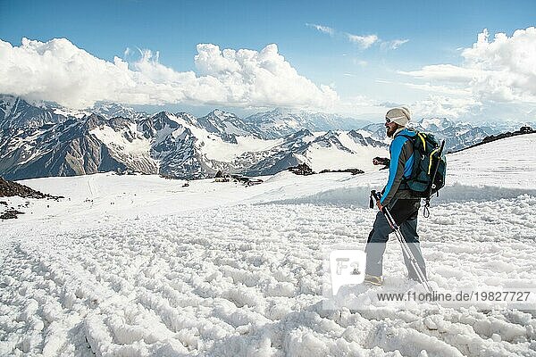 Müder Reisender mit Sonnenbrille und einem Rucksack  auf dem der Eispickel von einem schneebedeckten Gipfel herabhängt  vor dem Hintergrund schneebedeckter Berge und mit einem Stock für skandinavisches Wandern