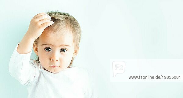 Kaukasische Baby Mädchen gegen helle weiße Wand  natürliches Licht. Porträt eines Kindes  das nach seiner Haarspange greift und in die Kamera schaut. Platz zum Kopieren auf der rechten Seite