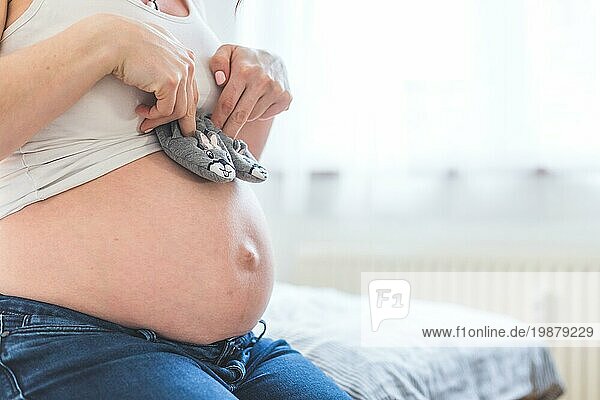 Caucasian schwangere Mutter hält Baby Schuhe in ihren Händen  nackten Bauch in der unscharfen Hintergrund