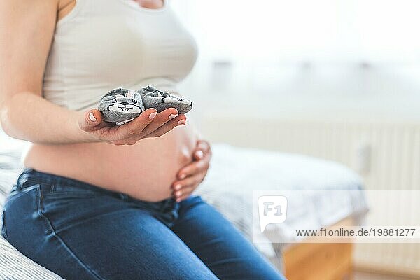 Caucasian schwangere Mutter hält Baby Schuhe in ihren Händen  nackten Bauch in der unscharfen Hintergrund
