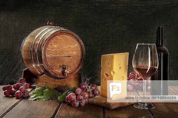 Ein Glas Rotwein mit einer Flasche  ein Weinfass  ein großes Stück Käse  Trauben und Weinblätter  Seitenansicht auf einem dunklen rustikalen Hintergrund  Low Key Foto mit einem Platz für Text