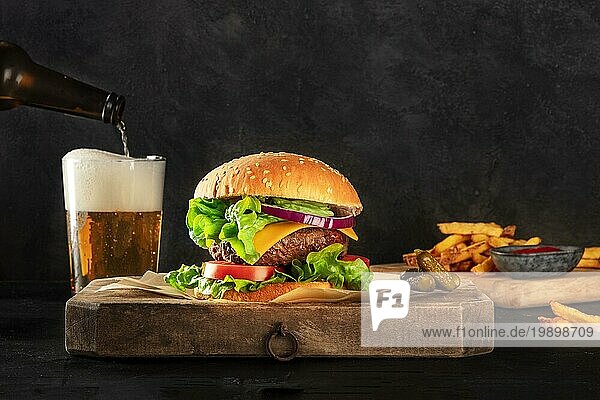 Burger und Bier. Hamburger mit Rindfleisch  Käse  Zwiebel  Tomate und grünem Salat  eine Seitenansicht auf einem dunklen Hintergrund mit Kopierraum  Gewürzgurken  Pommes frites und Ketchup. Selektiver Fokus