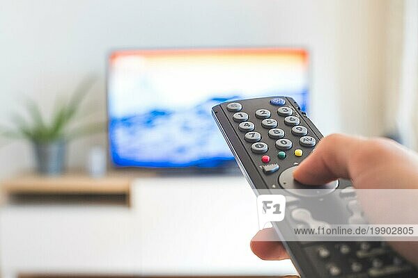 Eine TV Fernbedienung in der Hand  im Vordergrund  der Fernseher im verschwommenen Hintergrund. Streaming