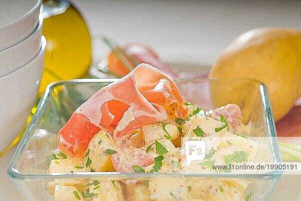 Frischer hausgemachter Parmaschinken und Kartoffelsalat  mit rohen Zutaten auf einem Tisch mit Schüsseln und Geschirr  Foodfotografie