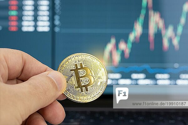 Hand hält eine Bitcoinmünze und Chart auf Laptop Display im Hintergrund. Cryptocurrency Handel oder Investitionen Konzept
