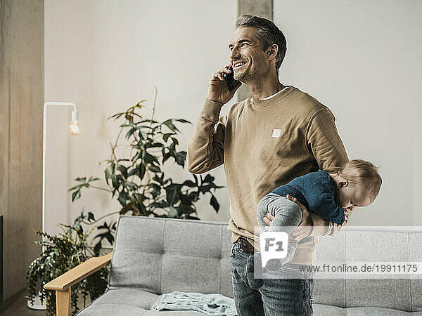 Smiling father holding baby boy and talking on smart phone at home