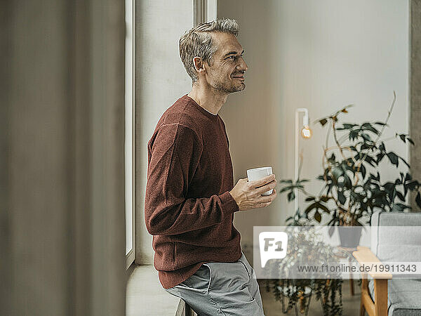 Smiling man with coffee cup leaning on windowsill at home