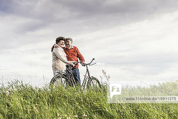 Happy senior couple standing with bicycle at field under cloudy sky