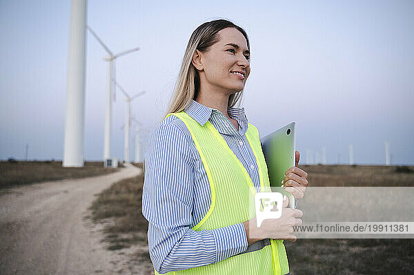 Smiling engineer holding laptop and standing in front of wind turbines