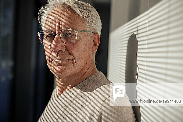 Smiling senior man wearing eyeglasses with sunlight over face near wall