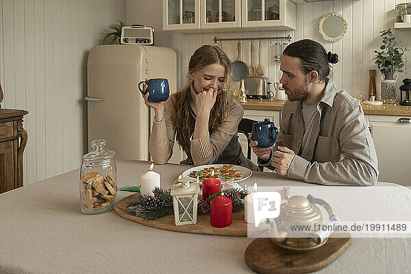 Smiling couple holding mugs with plate of cookies on table in kitchen