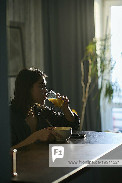 Young woman drinking orange juice and having breakfast in morning at home