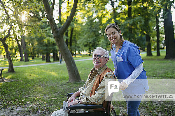 Smiling healthcare worker walking with senior man sitting in wheelchair near trees at park