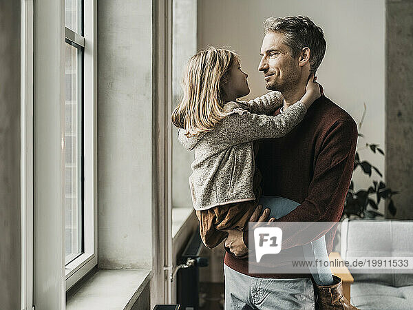 Smiling father holding daughter in arms near window at home